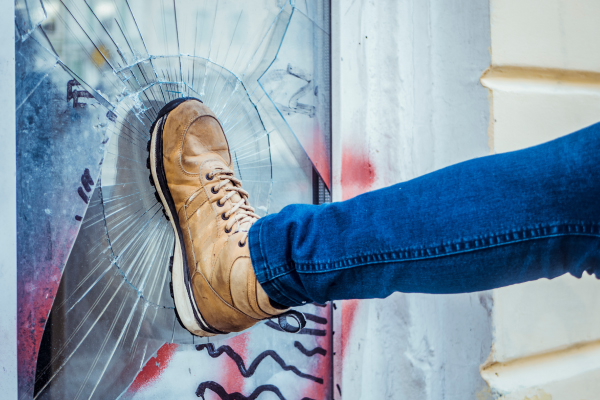 A brown boot kicks and breaks a window with scarlet and gray graffiti.
